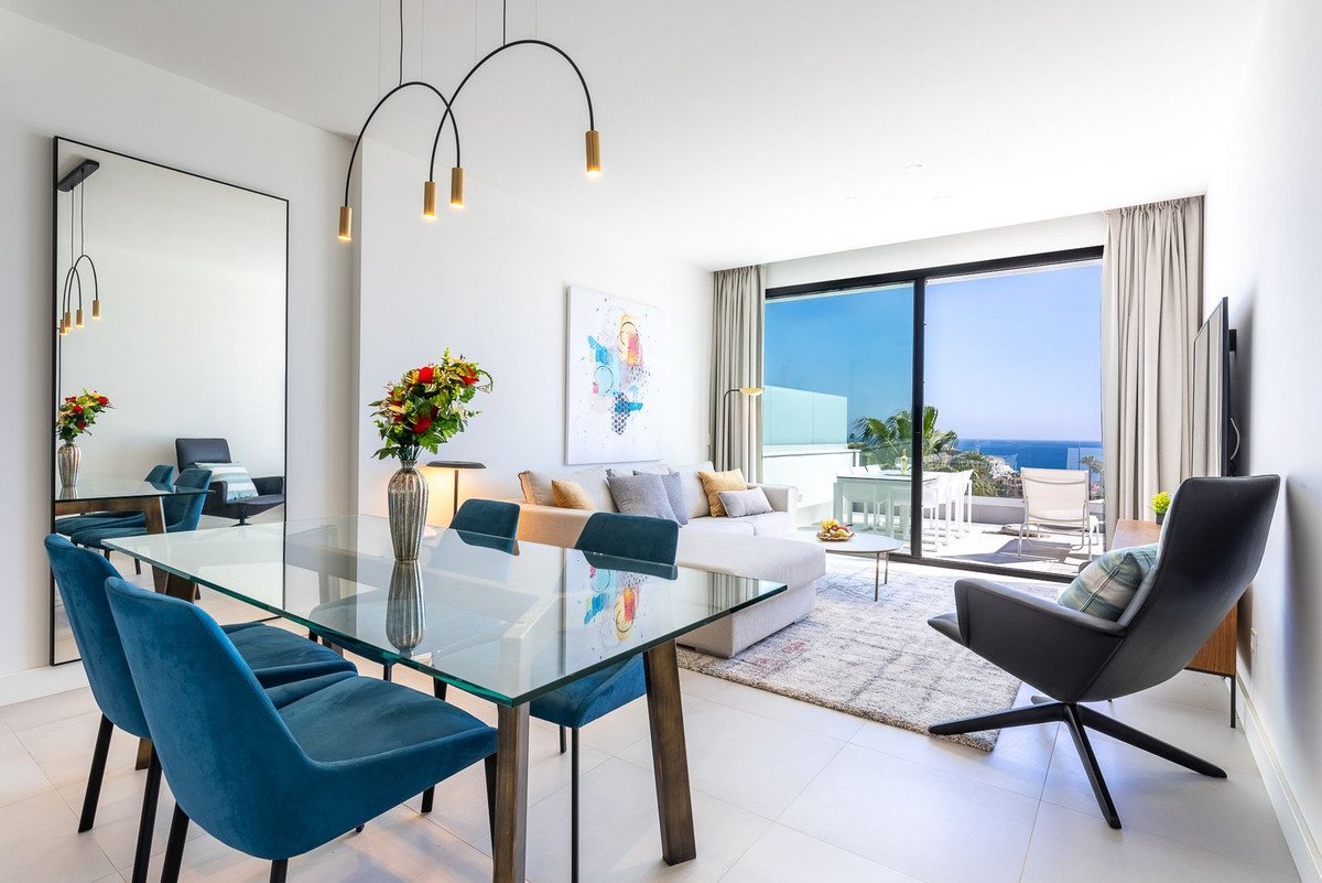 Modern living and dining area with blue chairs, a glass table, comfortable sofa, and balcony with sea view.
