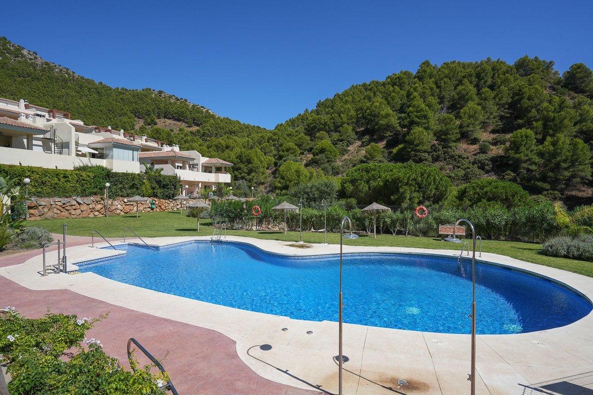 A resort-style swimming pool is set against a backdrop of lush green hills and residential buildings on a sunny day.