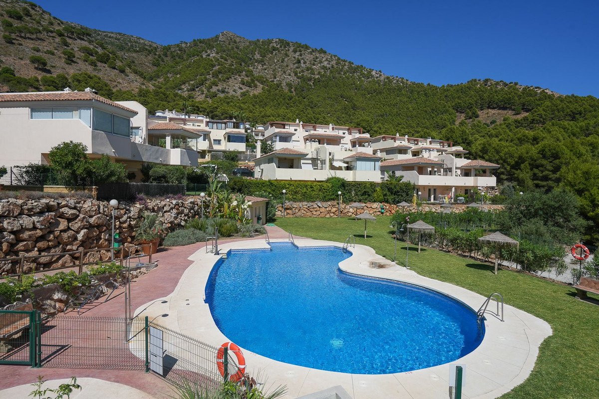 A bright blue swimming pool in front of a row of houses set against a lush green mountain and clear blue sky.