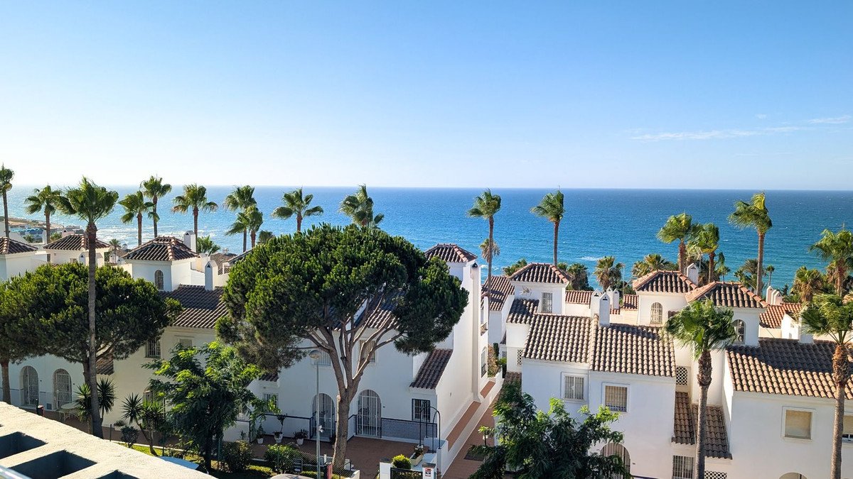 View of white buildings with terracotta roofs and palm trees, overlooking the blue sea under a clear sky.