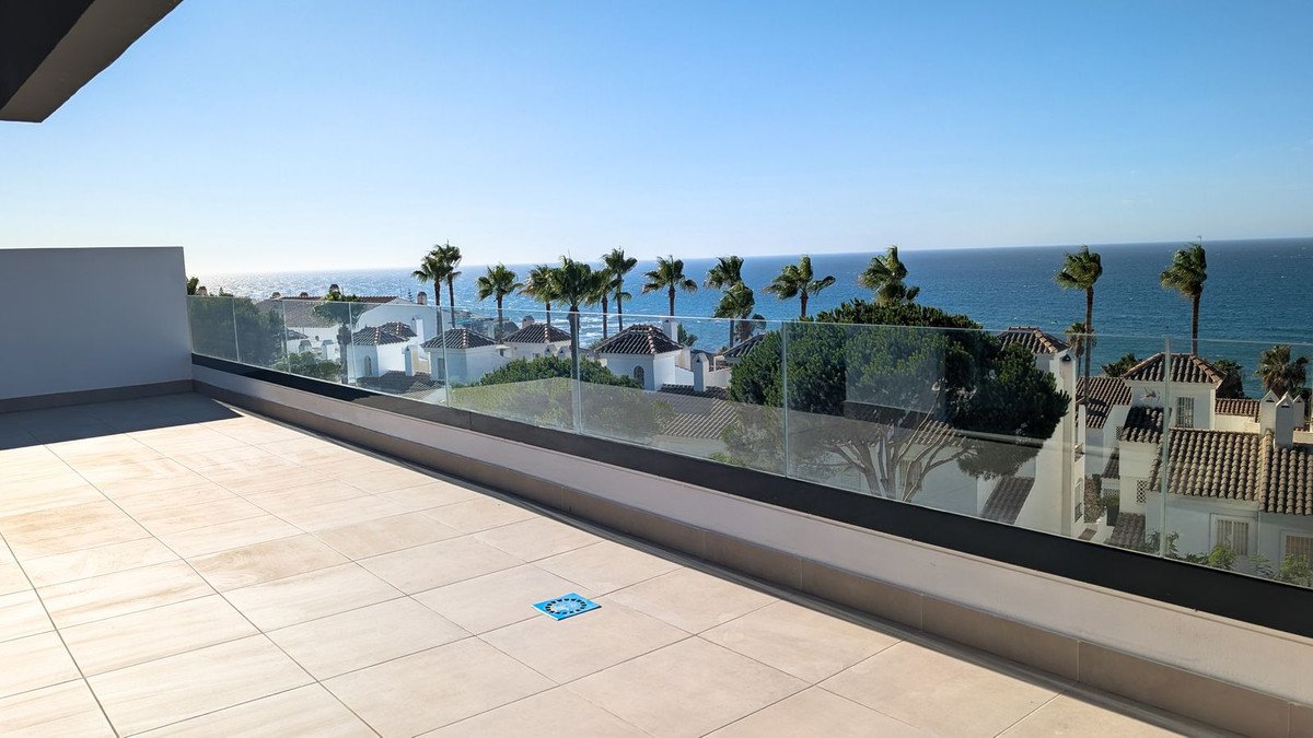 A spacious terrace with a glass railing offering a beautiful sea view, palm trees, and white houses below.