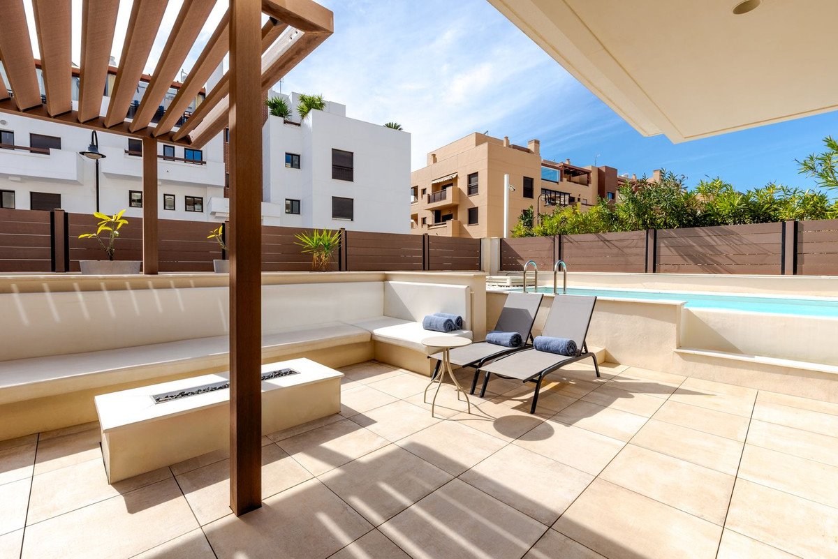 A modern terrace with a pool, lounge chairs, built-in seating, and a fire pit, under a wooden pergola.
