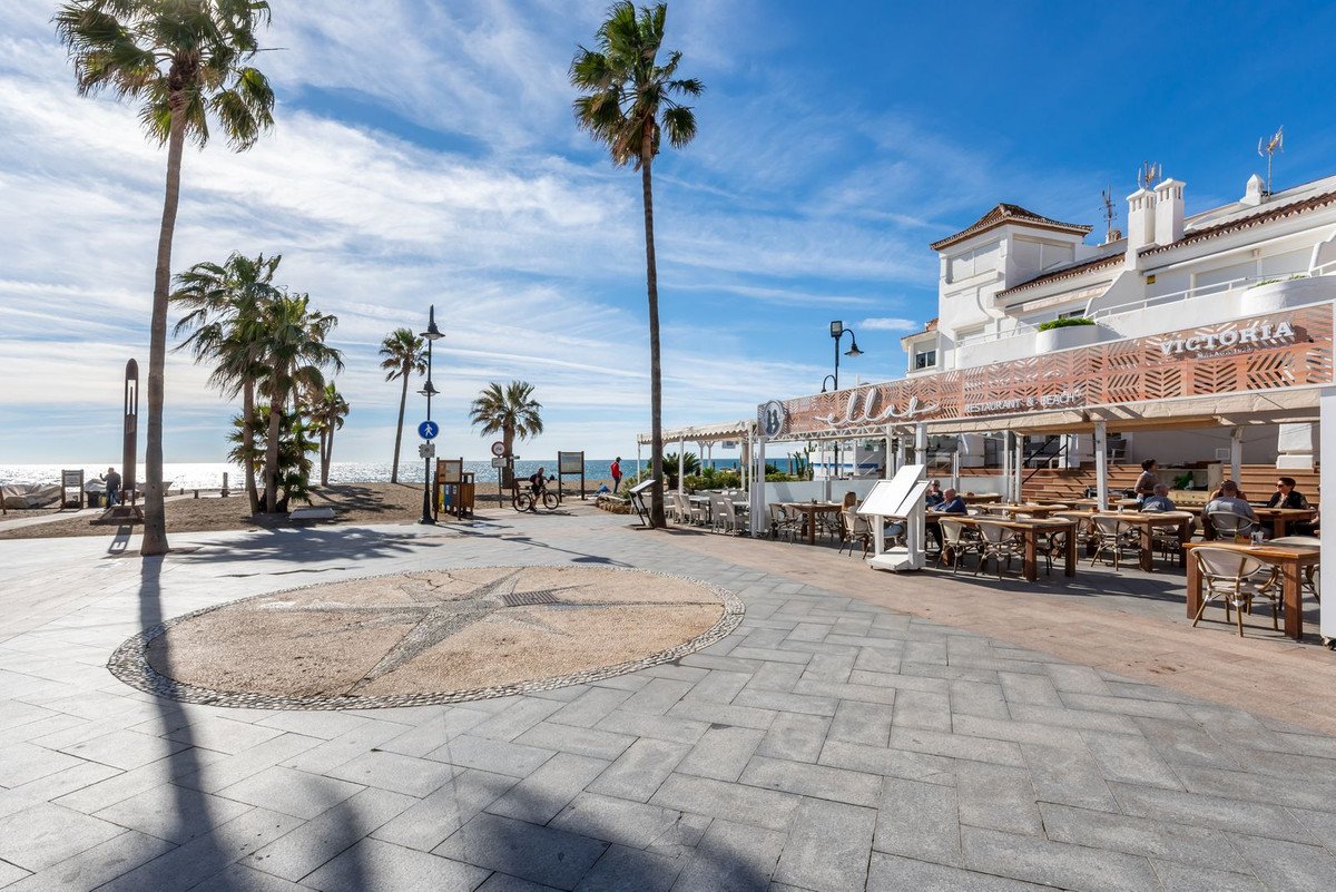 Outdoor seating area of a beachfront restaurant with palm trees and the sea visible in the background.