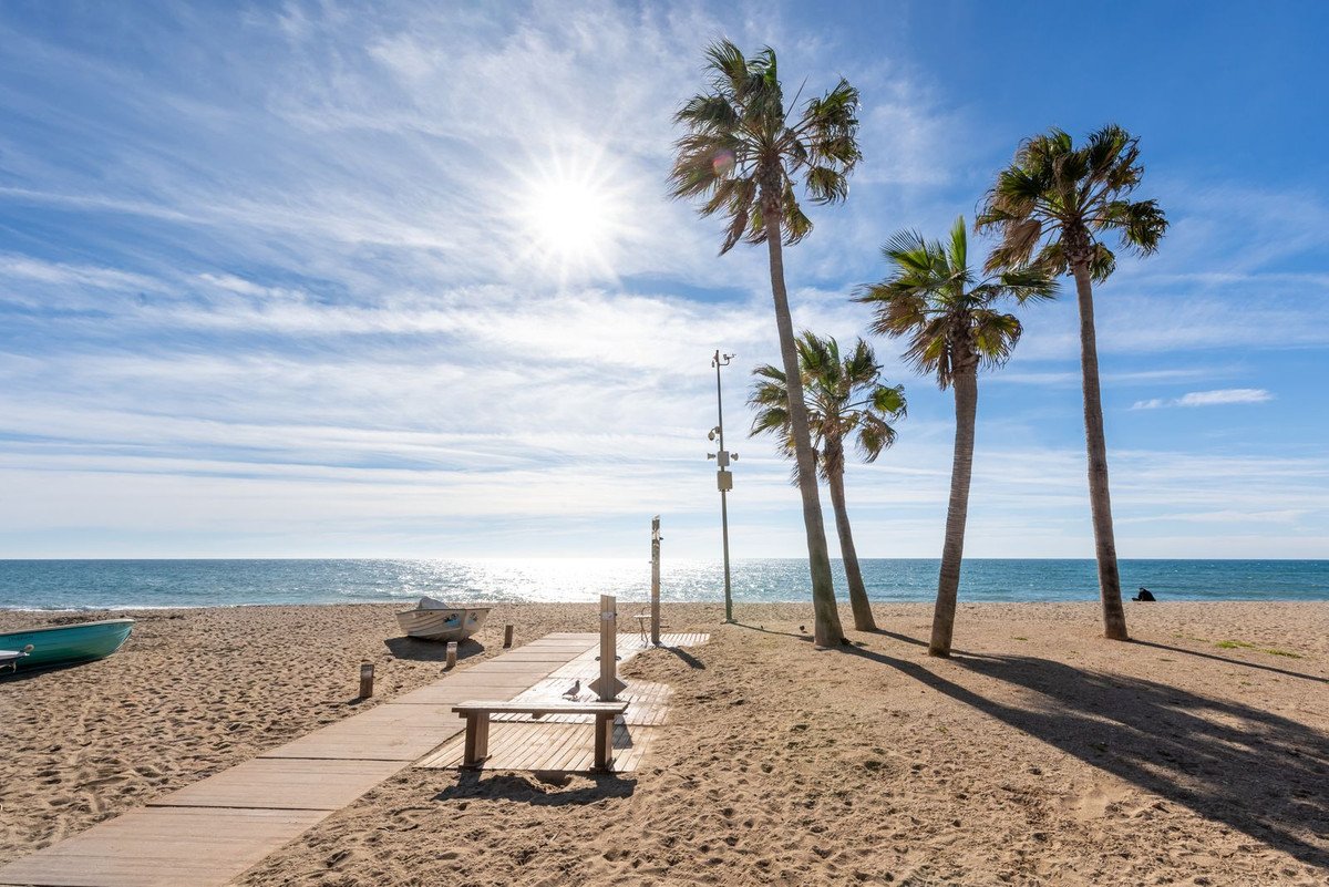 Sandy beach with palm trees, wooden walkway, and calm ocean under a bright sunny sky with wispy clouds.