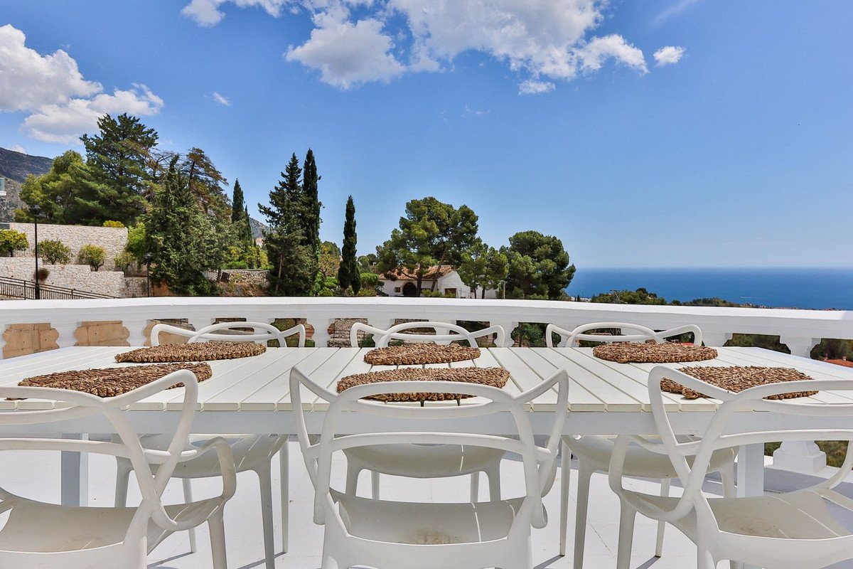White outdoor dining table and chairs on a terrace with a beautiful sea view and lush greenery.
