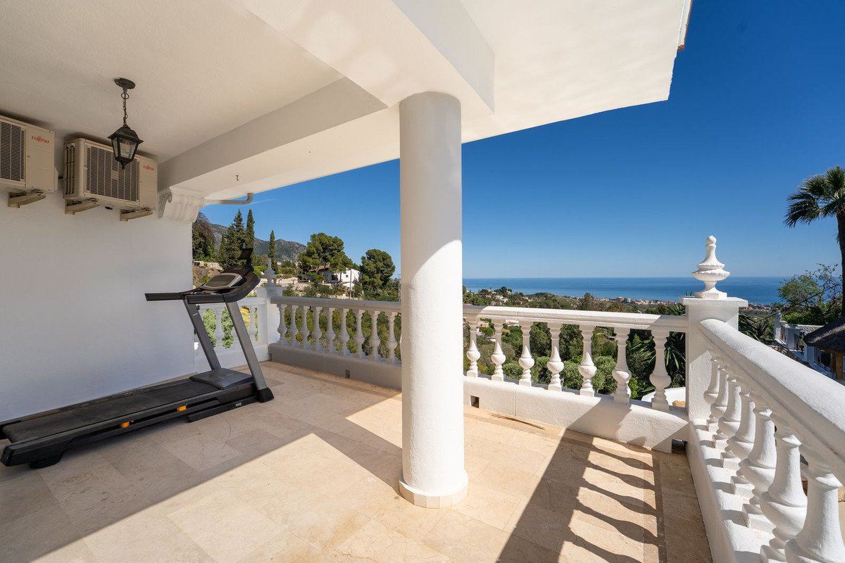 A spacious balcony with a treadmill and ornate railing offering a panoramic sea view and distant mountains under a clear blue sky.