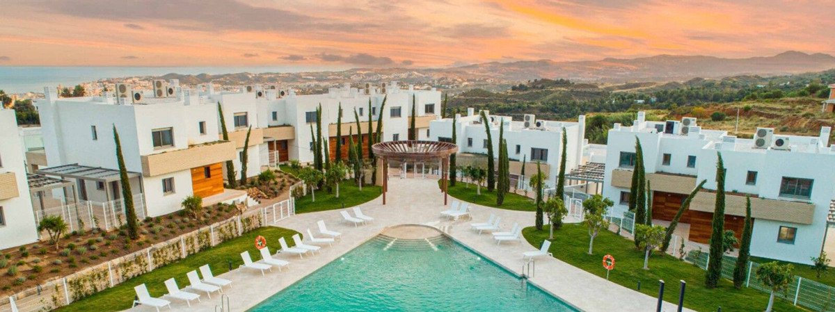 Modern white villas surrounding a swimming pool and lounge area, with a scenic hilly landscape and sunset sky in the background.