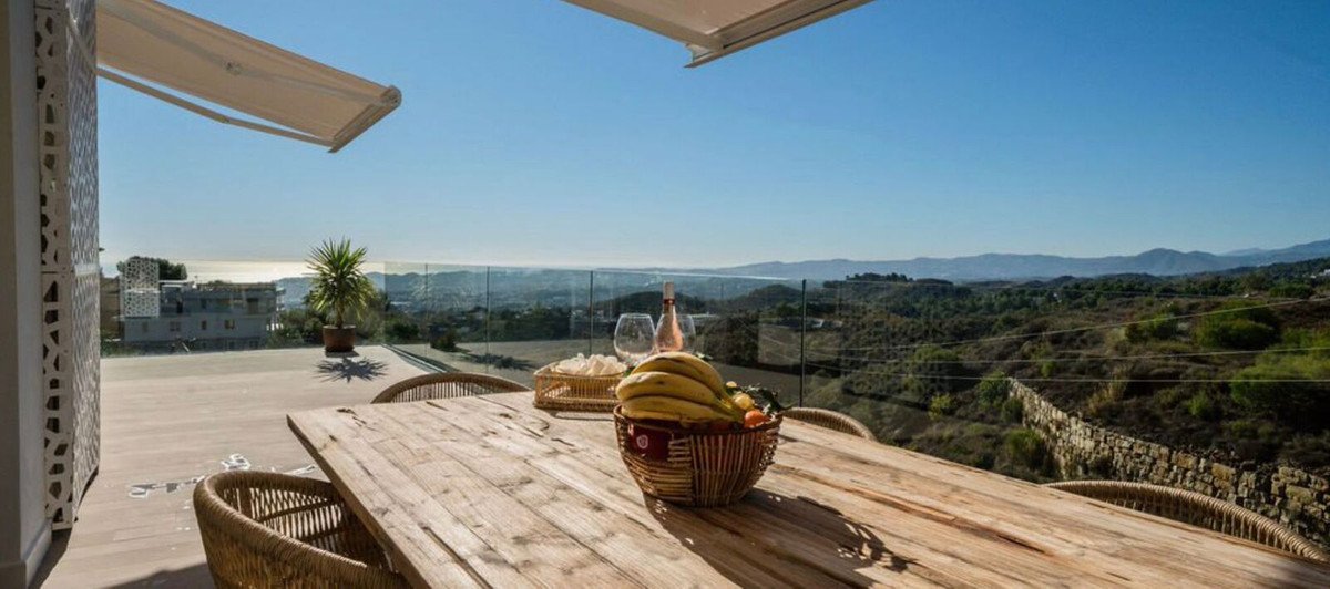 Outdoor terrace dining table with sea and mountain views, featuring a bottle of wine and fruit basket.