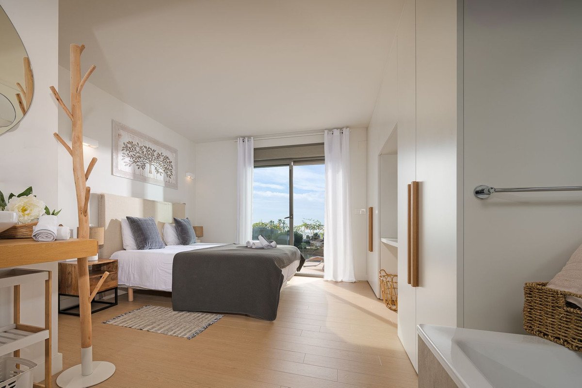 A stylish bedroom with a large bed, white curtains, and a glass door leading to a balcony with an ocean view.
