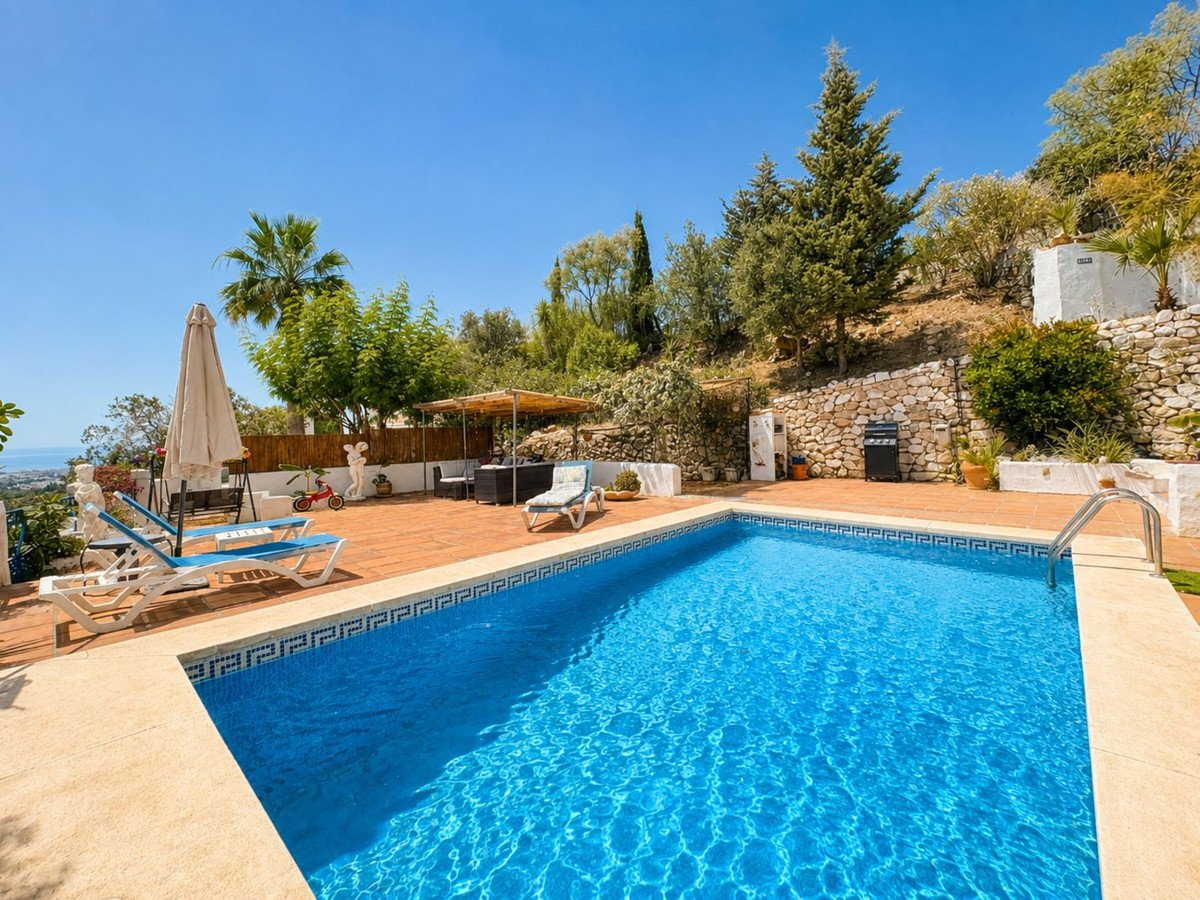 An inviting swimming pool with loungers, umbrellas, and a BBQ area, set against a hillside with distant sea views.