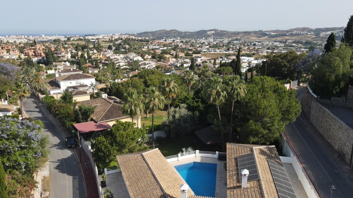 Aerial view of a villa with a swimming pool, surrounded by palm trees and overlooking a town and the sea.