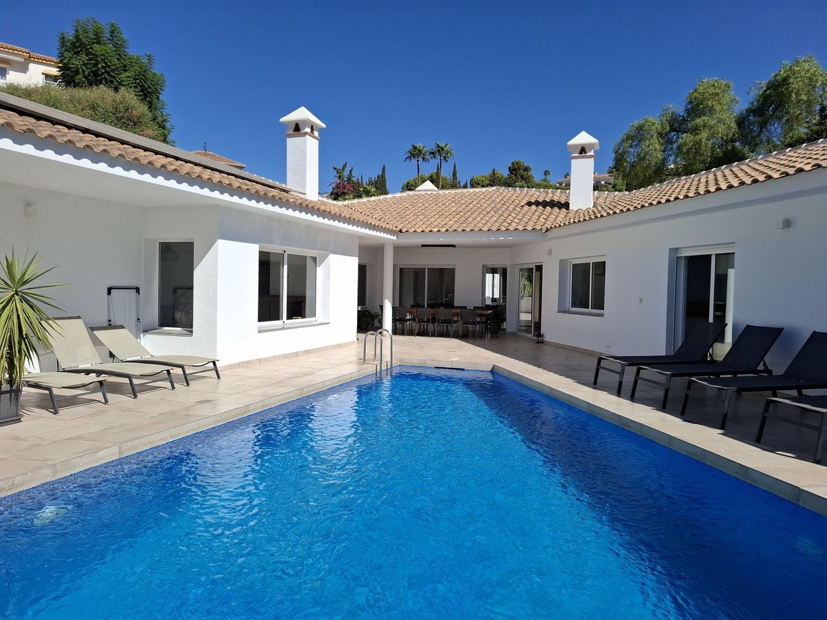 A modern villa features a large blue swimming pool, lounge chairs, and a covered patio with outdoor dining under a clear blue sky.