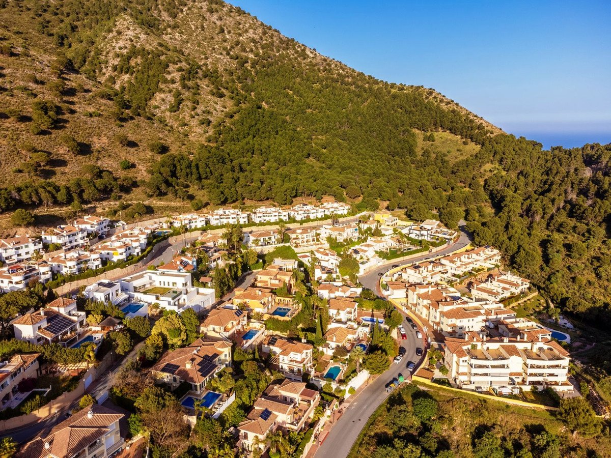 Aerial view of a hillside residential area with villas, pools, and lush greenery under a clear blue sky.