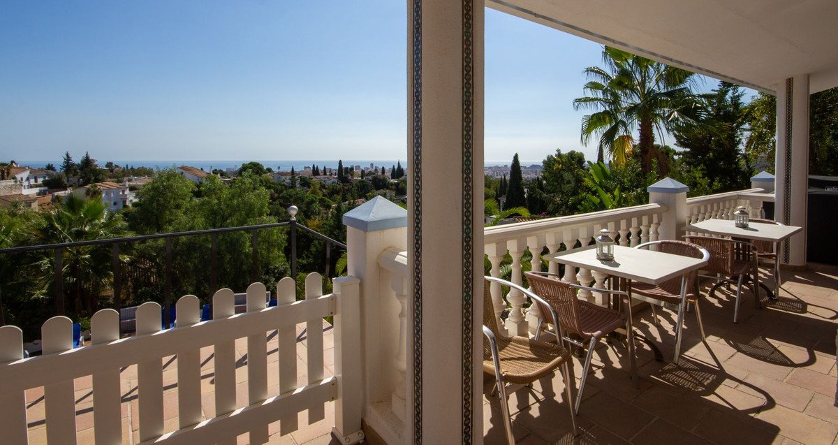 Terrace with dining tables and chairs, overlooking lush greenery and a distant sea view under a clear blue sky.