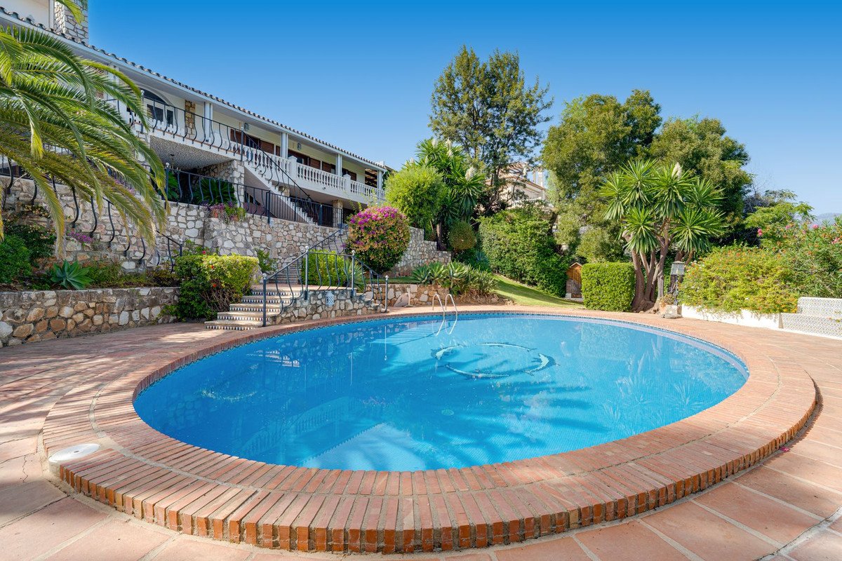Large oval swimming pool with a brick surround, overlooking a terraced villa surrounded by lush greenery and palm trees.