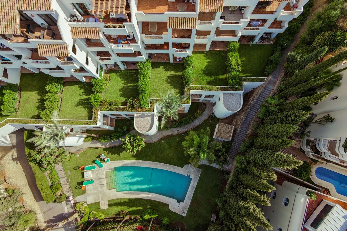 Aerial view of a white apartment building with balconies overlooking lush green gardens and a swimming pool.