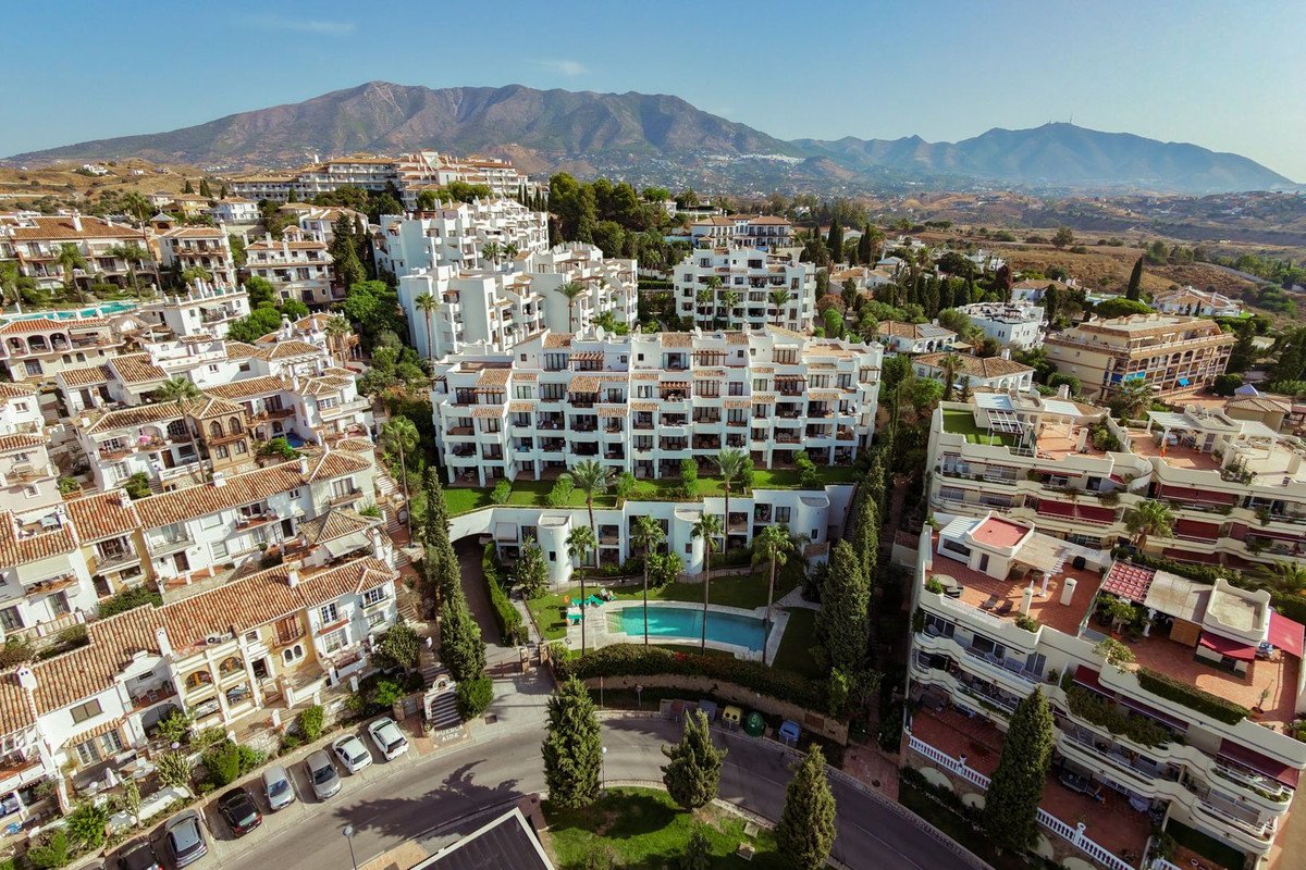 Aerial view of modern white apartment buildings with balconies, a swimming pool, and mountains in the background on a sunny day.