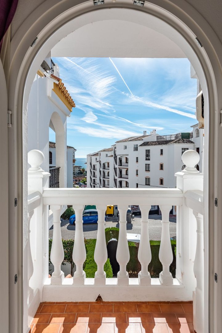 View from a balcony through an arched doorway overlooking white buildings, green lawns, street, and the sea in the distance.