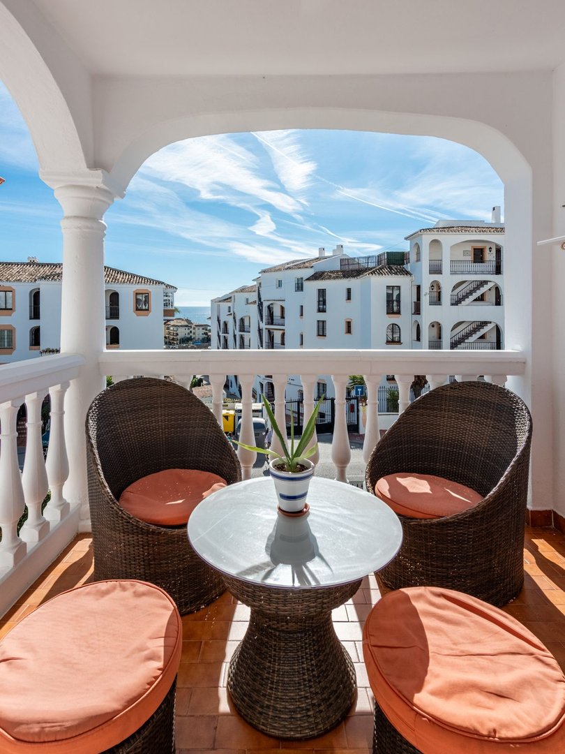 Charming balcony with rattan seating, a small table, and a view of white buildings and the sea under a blue sky.