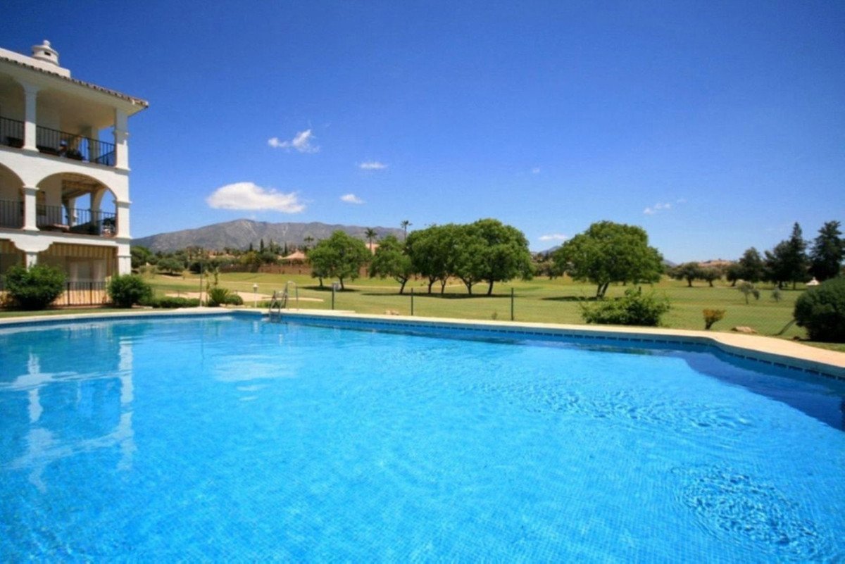 Apartment building with balconies next to a large blue swimming pool overlooking a green golf course with mountains in the background.