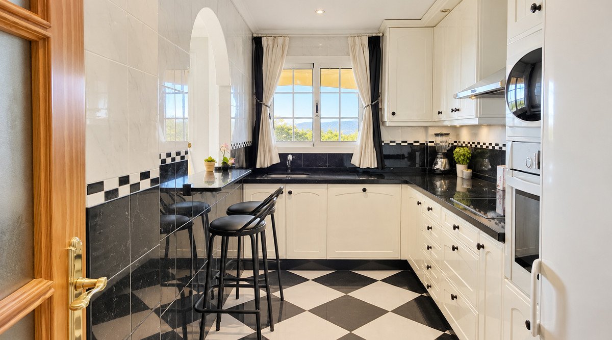 A kitchen with black and white checkered flooring, white cabinets, and a sea view through the window.