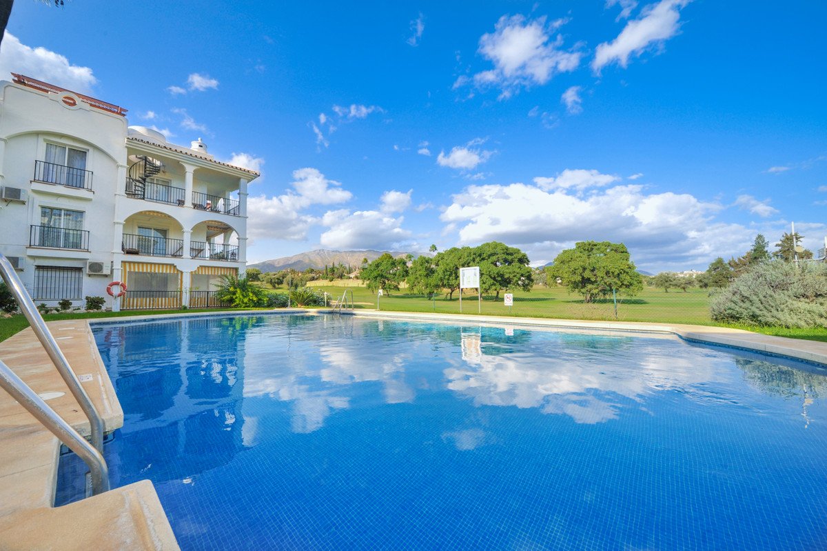 A communal swimming pool with a golf course and mountains in the background under a blue sky with clouds.