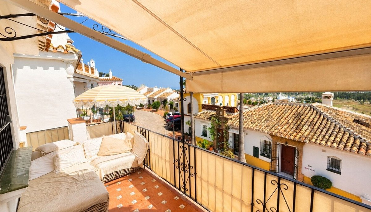 A furnished balcony with a lounge sofa and a striped umbrella overlooks a street with houses and a tiled roof landscape.