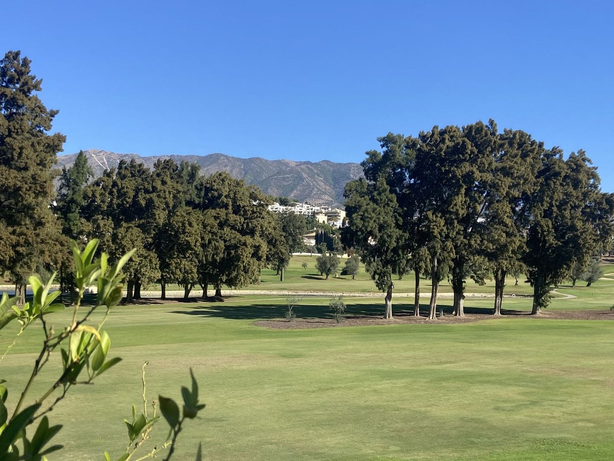 Expansive golf course with lush green fairways and trees, overlooking mountains and buildings under a clear blue sky.