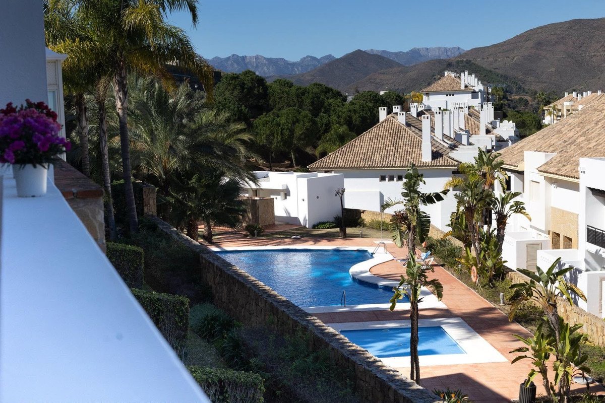 View from a balcony overlooking a swimming pool complex with palm trees and mountains in the distance. Sunny day.