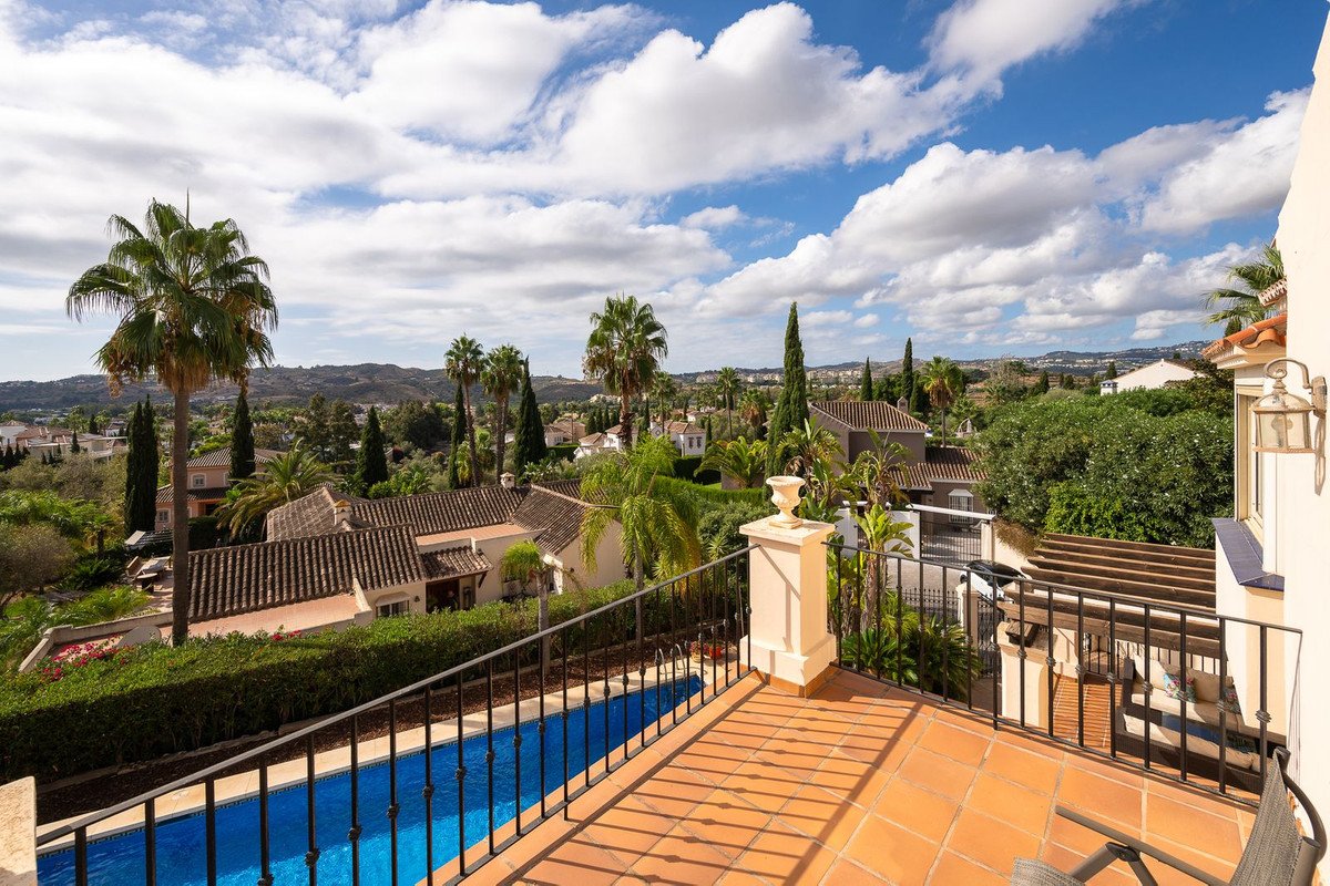 Terrace view overlooking a swimming pool, palm trees, and houses with a distant hilly landscape under a cloudy sky.