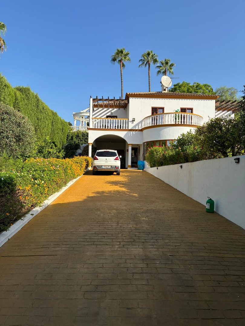 A white villa with palm trees and a curved balcony, accessed via a long paved driveway with lush green landscaping.