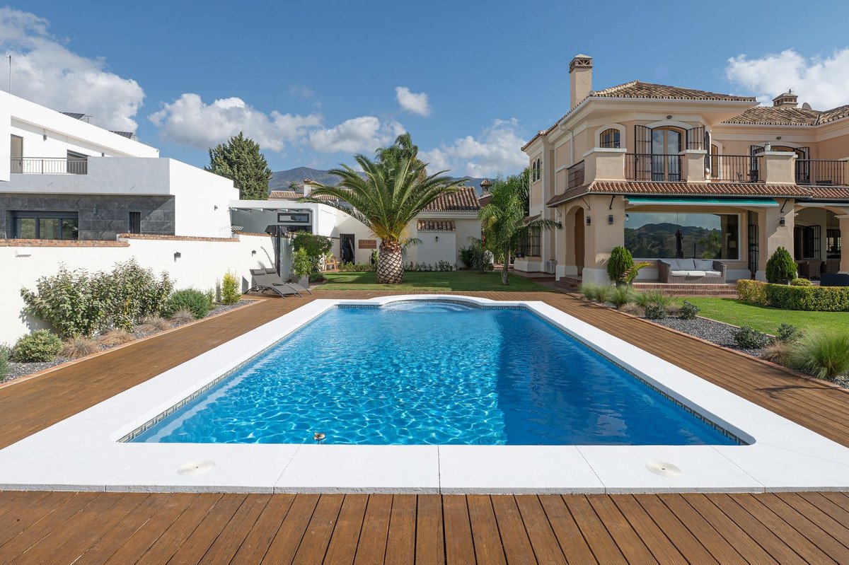 Luxurious villa with a swimming pool, manicured garden, palm tree, and mountain view under a blue sky.