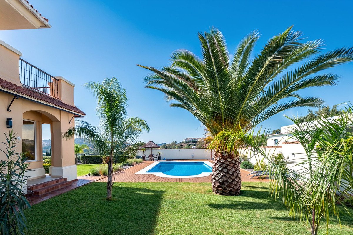 A luxury villa with a swimming pool, surrounded by palm trees, lush green lawn, and a wooden deck on a sunny day.