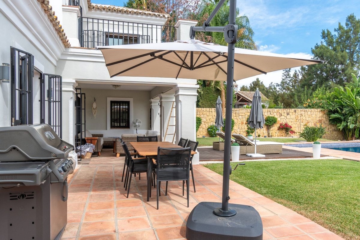 Outdoor dining area with a grill, large umbrella, and lounge chairs by the pool and garden.