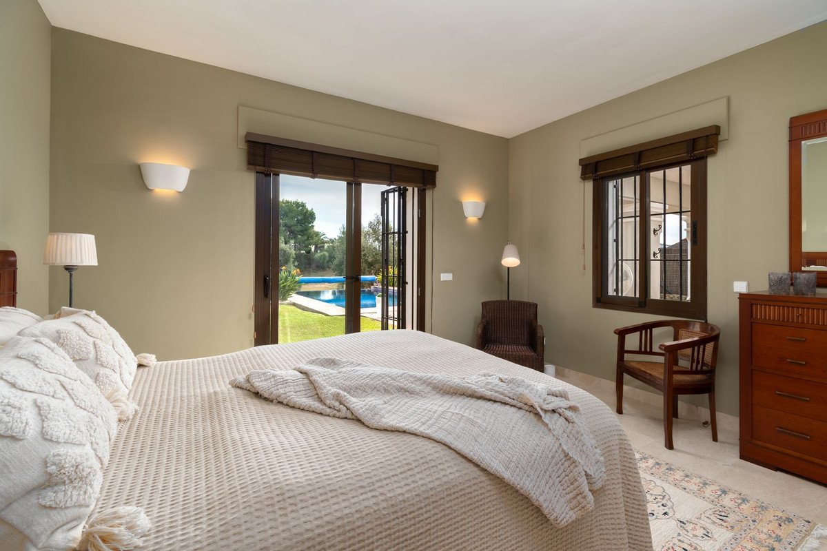 A bedroom with a large bed, doors opening to a garden with a swimming pool, and two chairs.