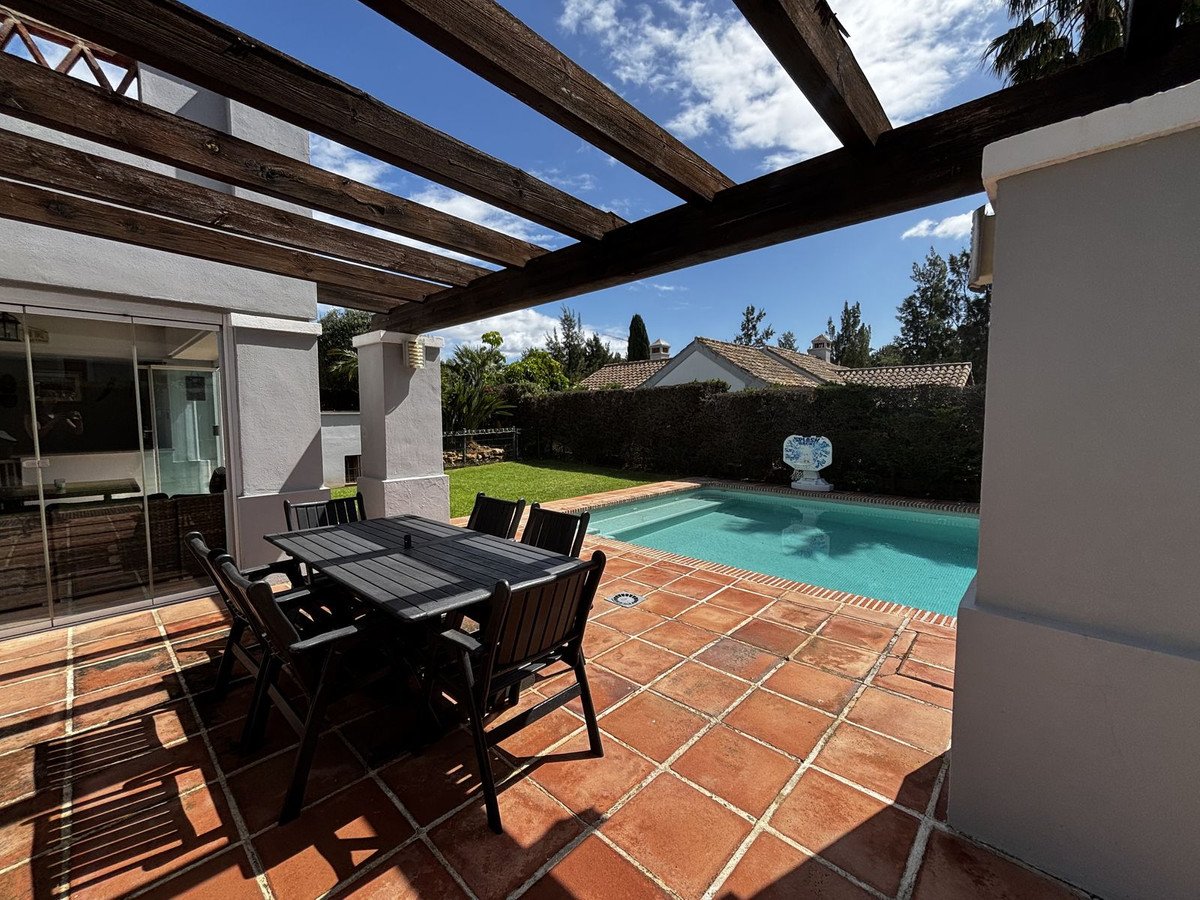 Covered patio with dining table, pool, and garden view under wooden pergola.