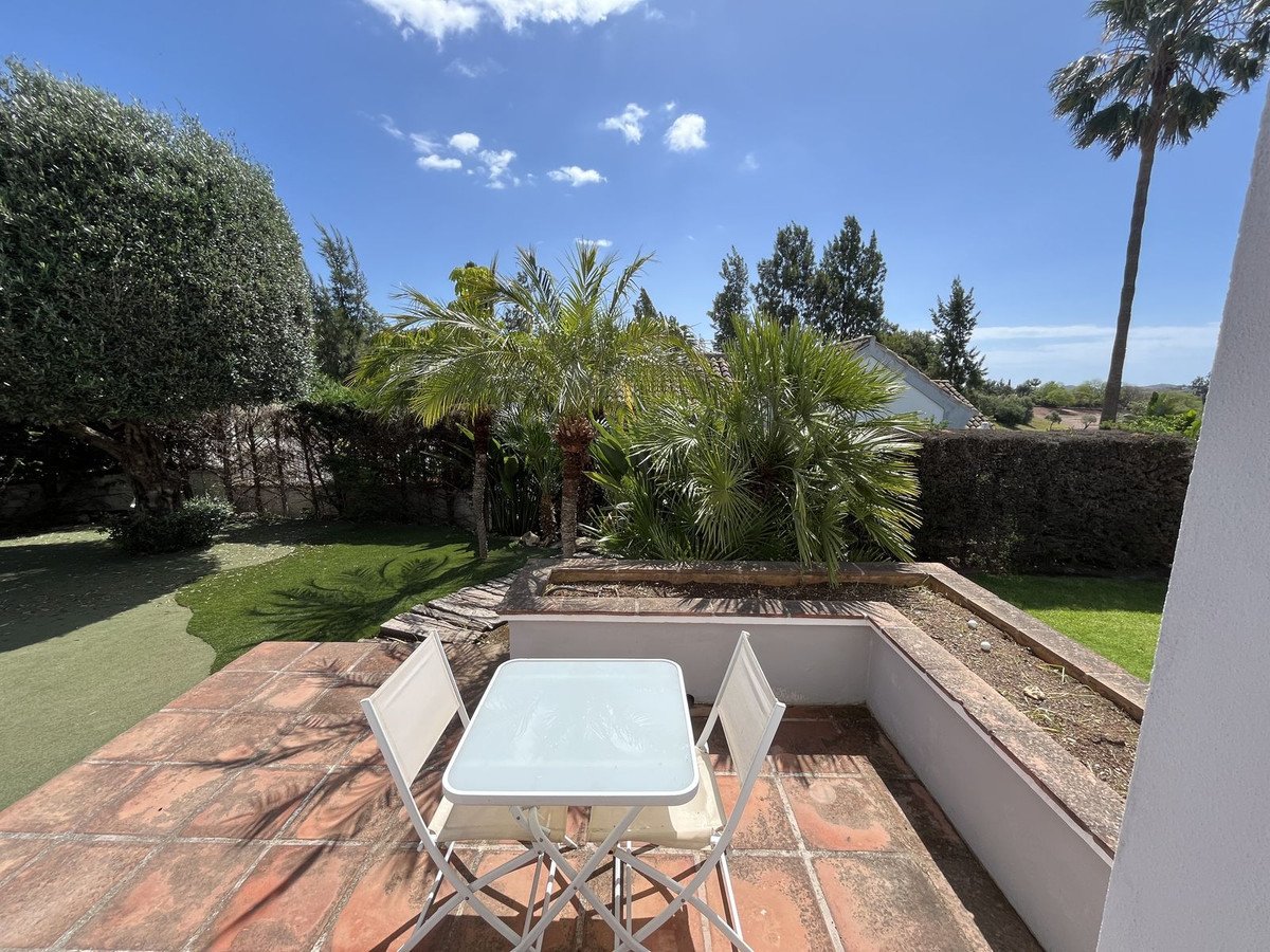 Terrace with white table and chairs, overlooking a lush garden with palm trees and green lawn under a blue sky.