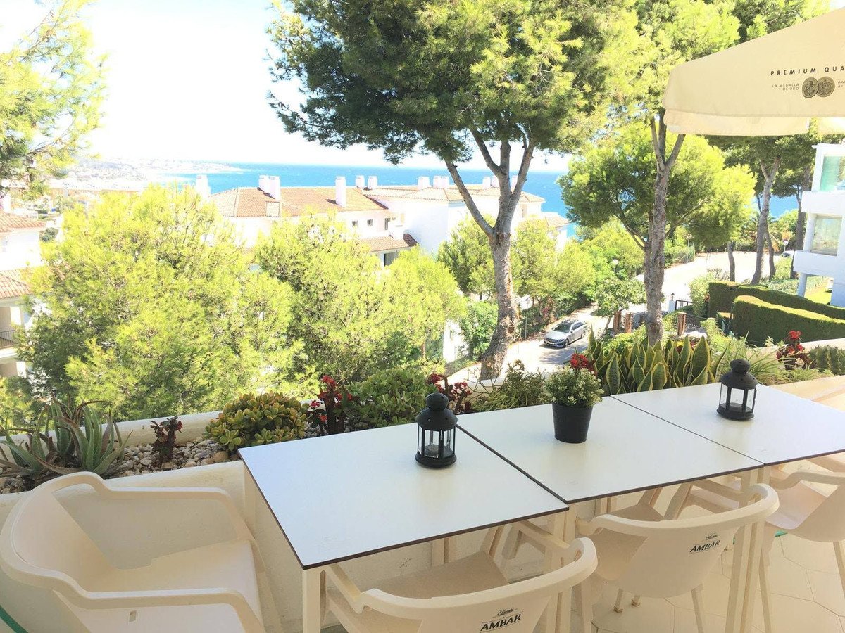 Terrace with two tables and chairs overlooking the sea, surrounded by lush greenery and trees.