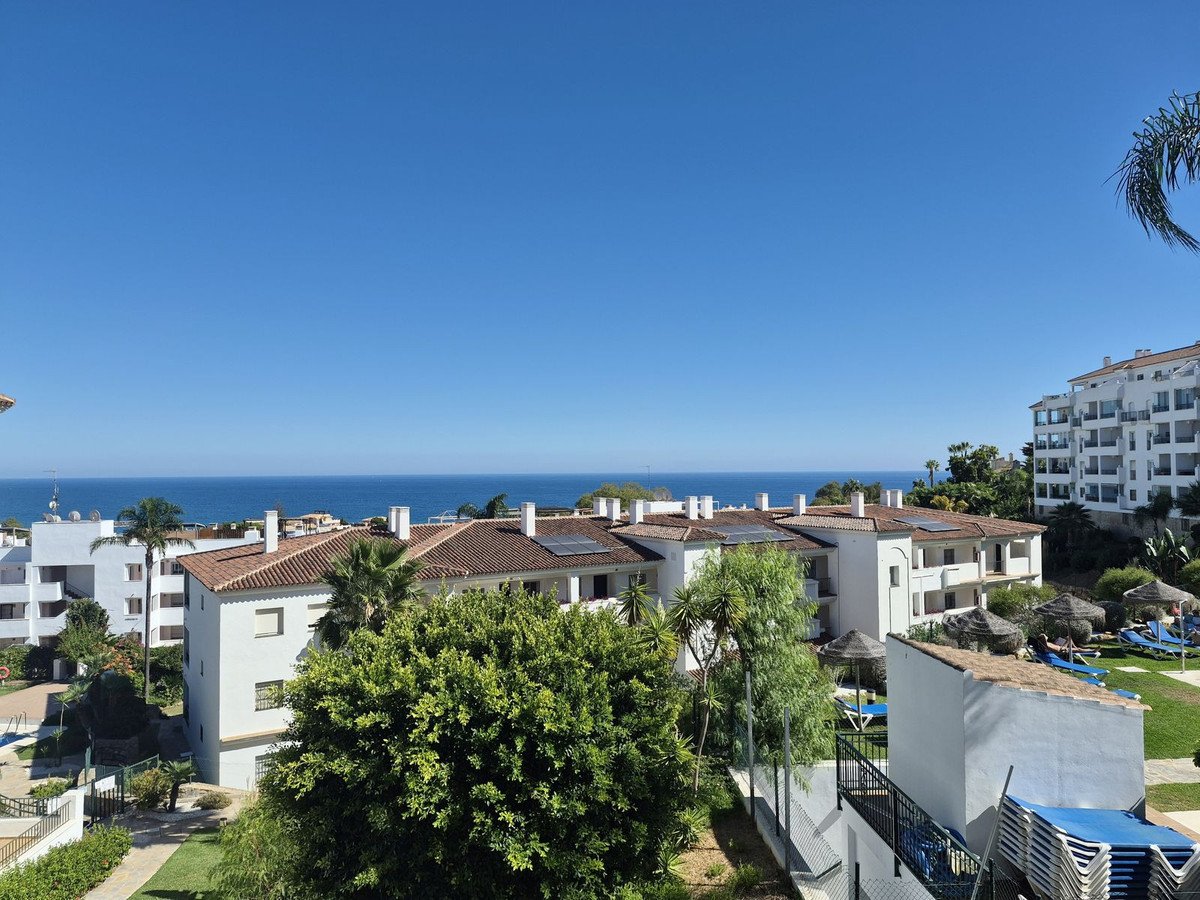 View of the sea from a balcony overlooking apartment buildings and a swimming pool area with sun loungers.