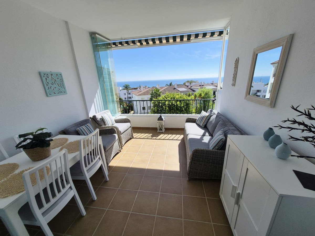 A bright balcony with dining table and seating area overlooking the sea with houses and trees.