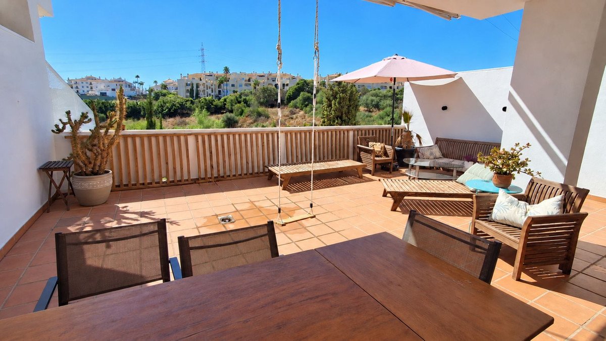Spacious terrace with wooden furniture, including a dining table, sofa, and a swing, overlooking a hillside with buildings.