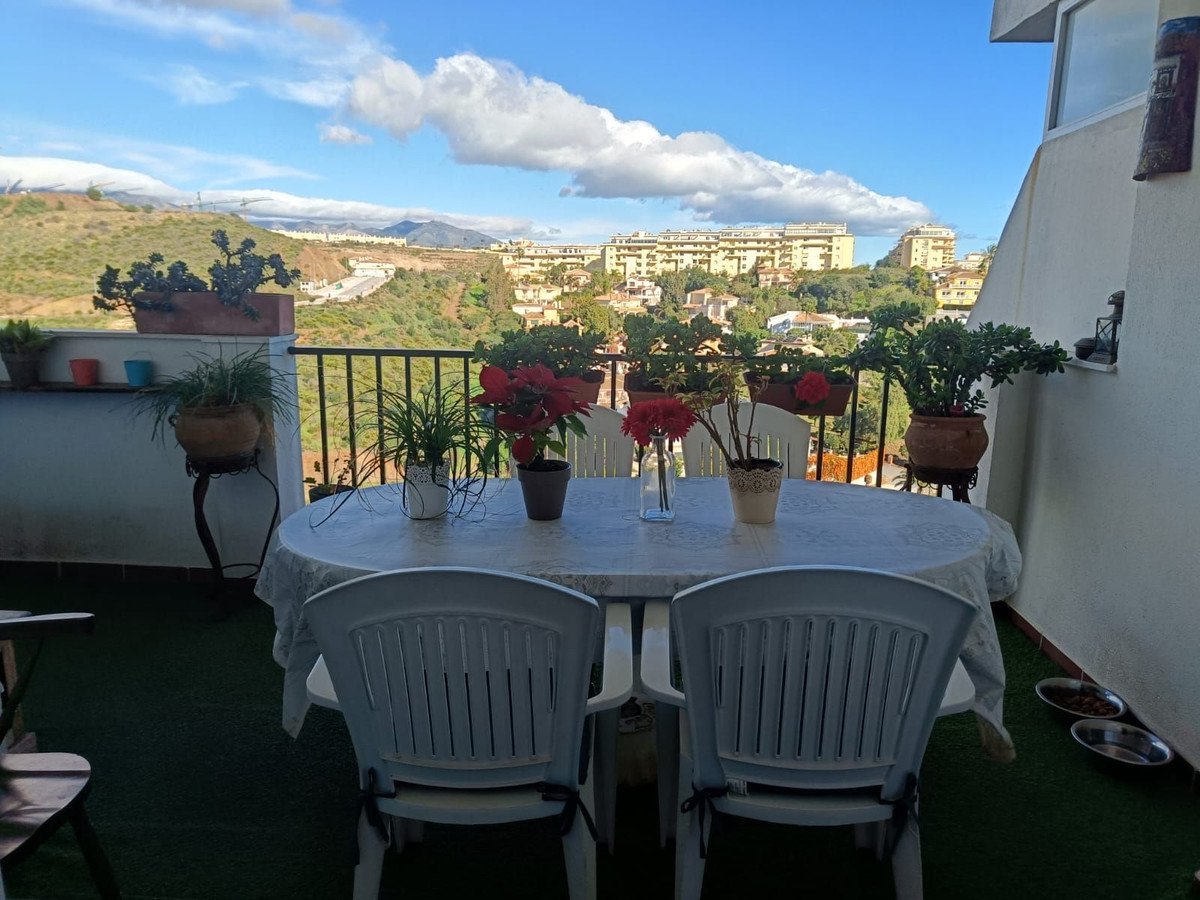 A balcony with a table and chairs, overlooking a town and hills under a cloudy sky.