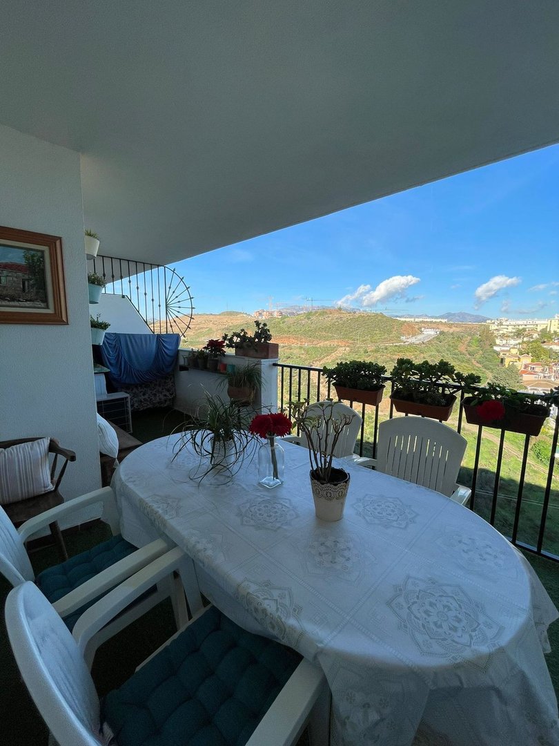 A furnished balcony with a table and chairs offers a view of green hills and a distant town under a blue sky.
