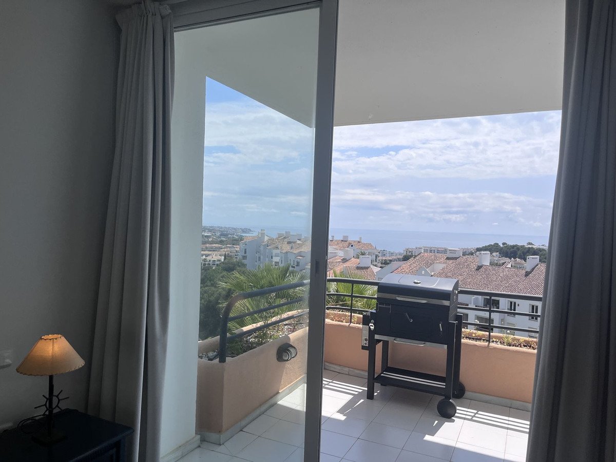 View from a room through a sliding door to a balcony with a grill, overlooking a town and the sea.