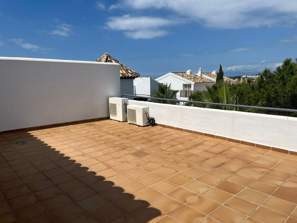 A spacious tiled terrace with two air conditioning units, overlooking rooftops and greenery under a blue sky.