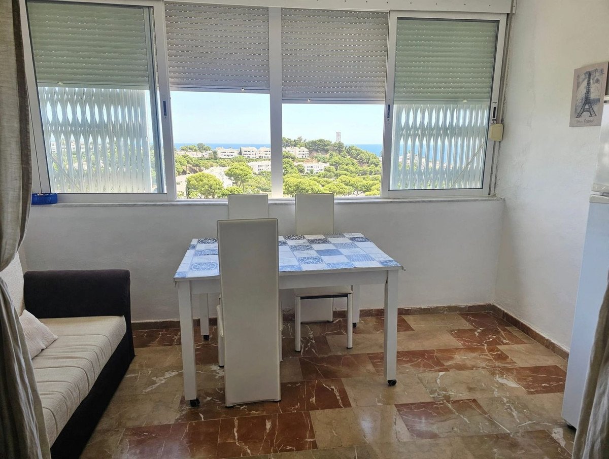A dining table with chairs and a sofa next to large windows with a view of the sea and trees.