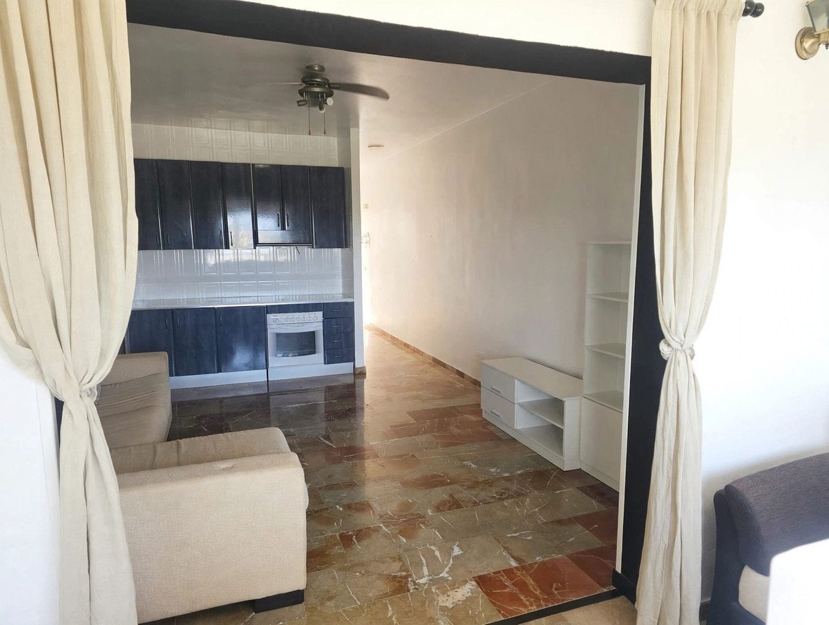 A view of a living area with beige sofas and a kitchenette featuring dark cabinets and a white oven. Marble flooring is visible.