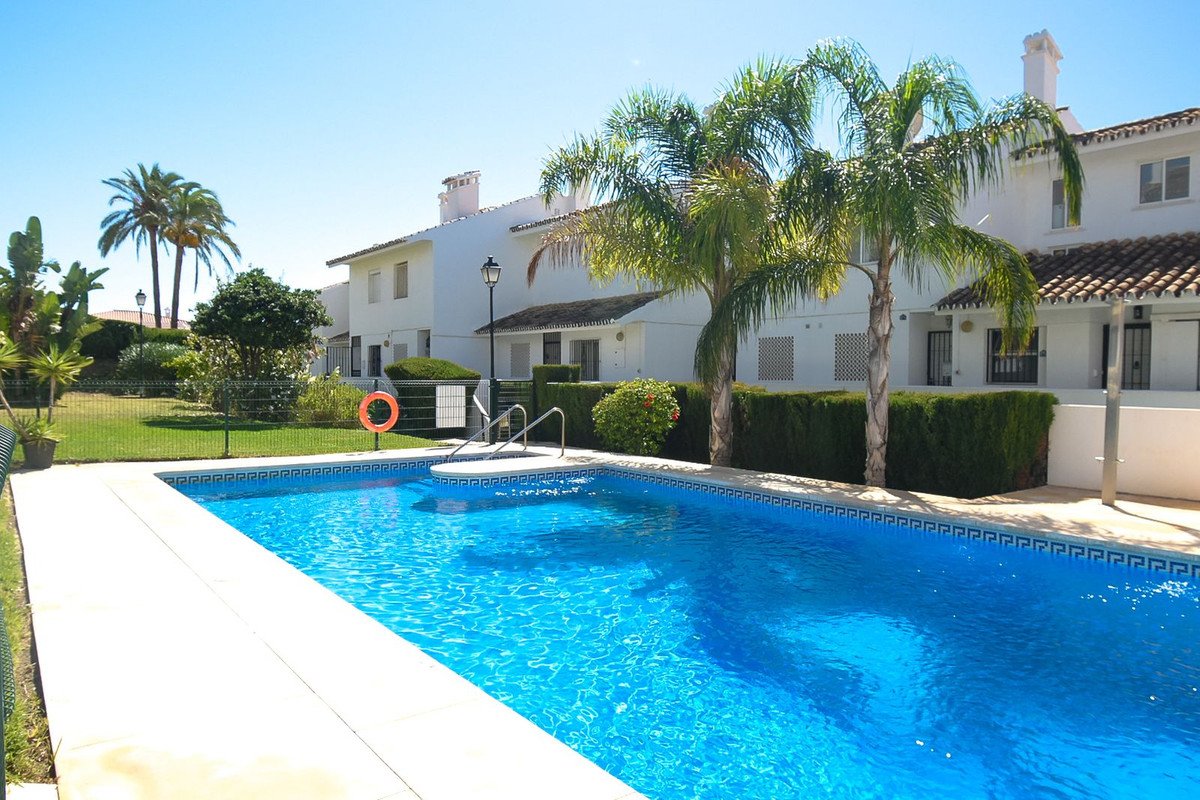 A bright blue swimming pool is in front of white townhouses, surrounded by palm trees and green grass.