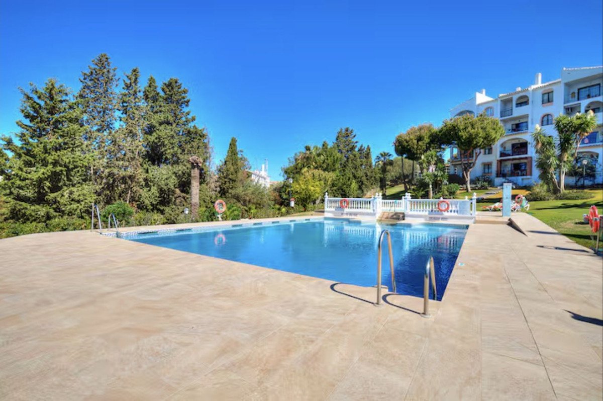 A bright blue swimming pool with tiled surroundings, surrounded by lush green trees and a white apartment building under a clear blue sky.