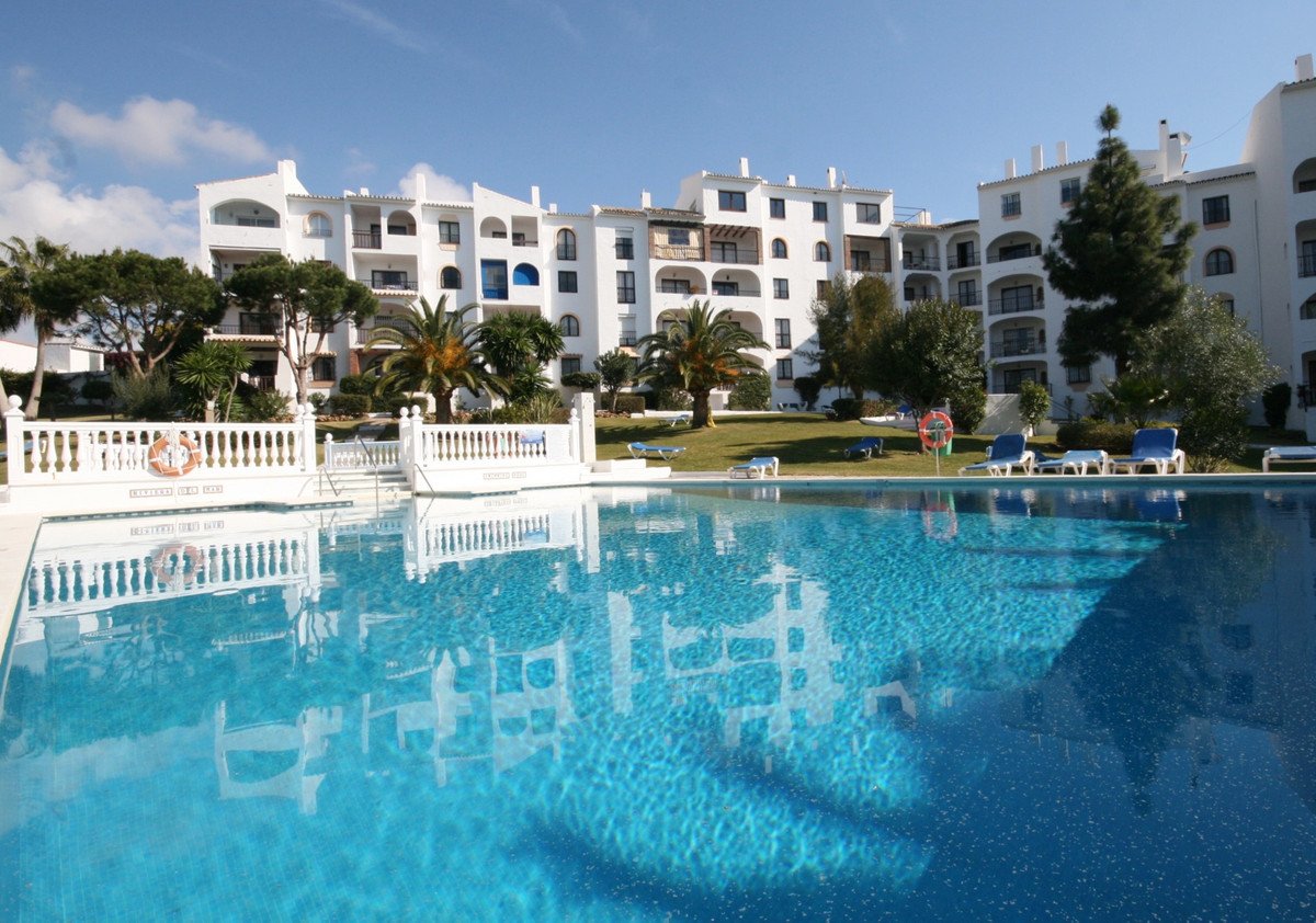 View of a large outdoor swimming pool with reflections of white apartment buildings under a blue sky.